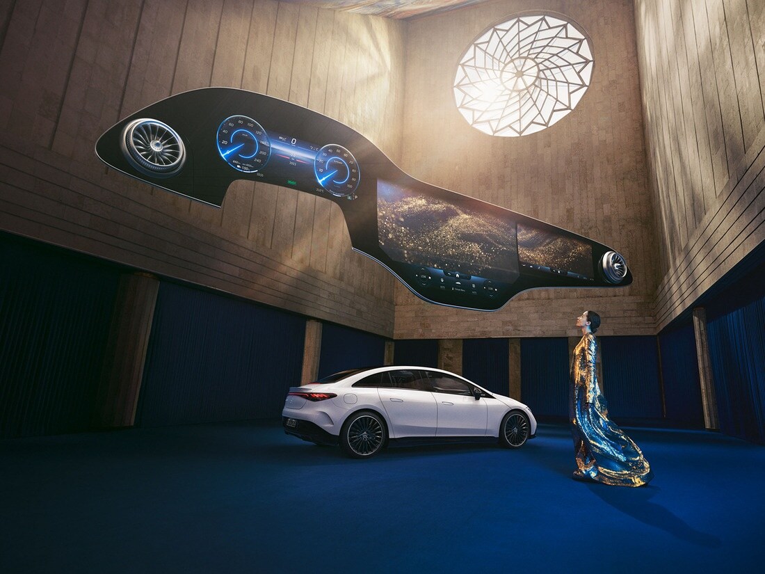 Kobieta stojąca przed elektrycznym Mercedesem z unoszącym się nad nią ekranem Hyperscreen. System multimedialny MBUX | Mercedes-Benz