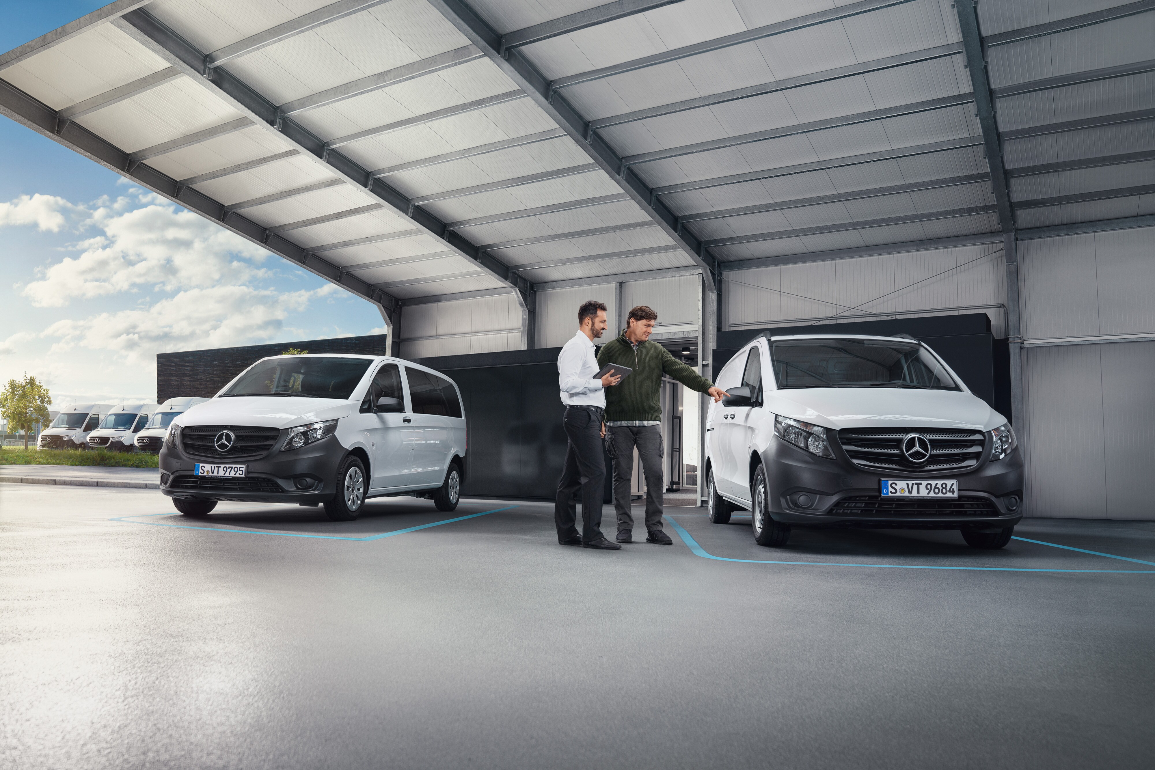 Wygoda | Samochody dostawcze Mercedes-Benz Dwie osoby rozmawiające w serwisie, stojąc przed dwoma białymi samochodami dostawczymi Mercedes-Benz.
