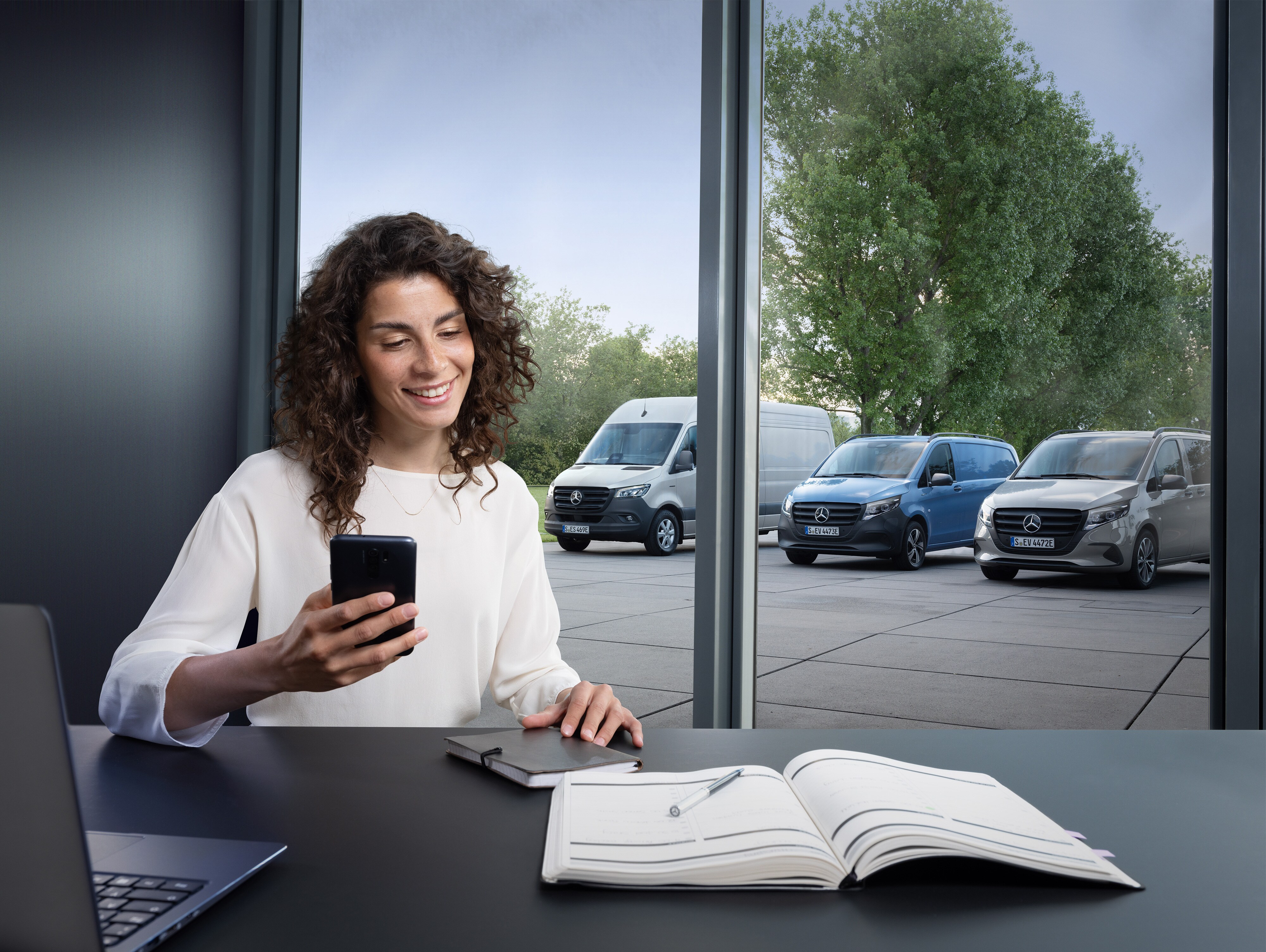 Mercedes-Benz | Wygoda | Rezerwacja wizyt online Kobieta ze smartfonem stoi przed oknem z trzema samochodami dostawczymi Mercedes-Benz widocznymi na zewnątrz.