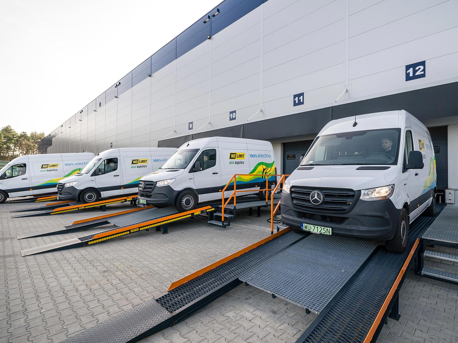 eSprintery w No Limit – idealny partner w miejskiej logistyce Cztery białe Mercedesy eSprinter na rampie przed magazynem firmy No Limit.